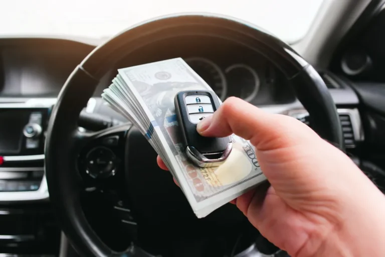 A hand holding a car key and US dollar banknotes inside a vehicle