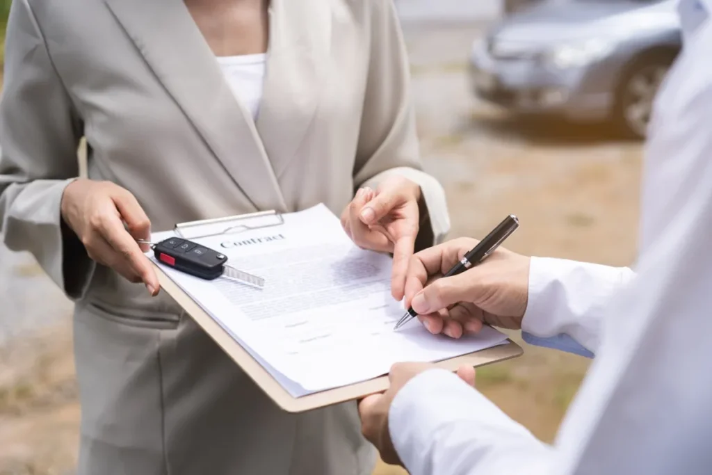 An agent handing a vehicle key to a buyer alongside signed contract paperwork